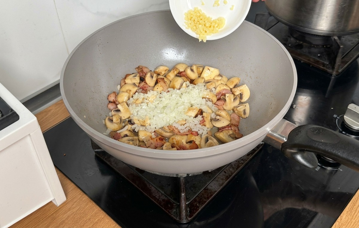 Vertiendo cebolla y ajo finamente picados desde un bol blanco sobre los champiñones y el beicon en el wok.