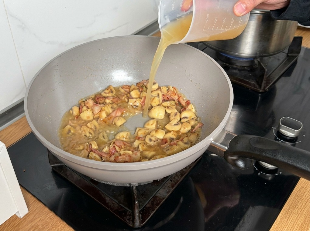 Caldo de pollo vertiéndose desde una jarra medidora a la mezcla de champiñones y beicon.