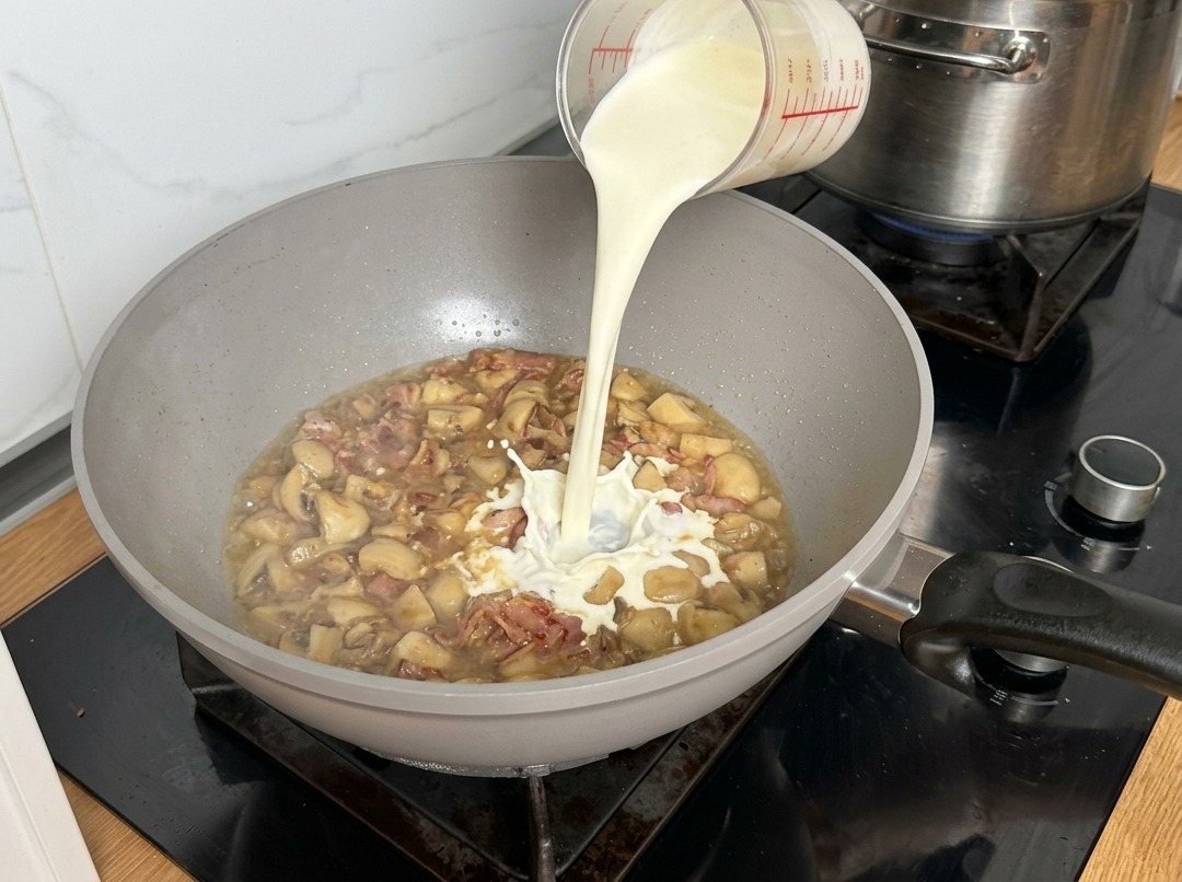 Crema de leche vertiéndose en la sartén, creando salpicaduras al contacto con la base de champiñones.