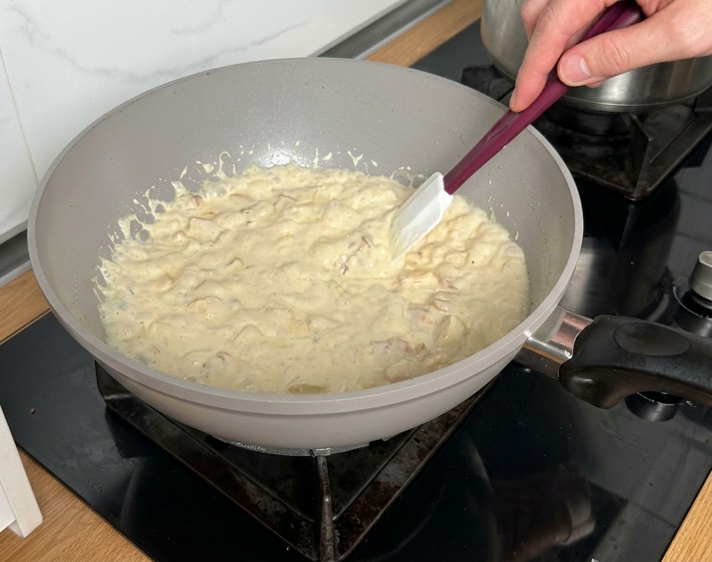 Una espátula blanca removiendo una salsa de crema espesa y burbujeante con beicon y champiñones.