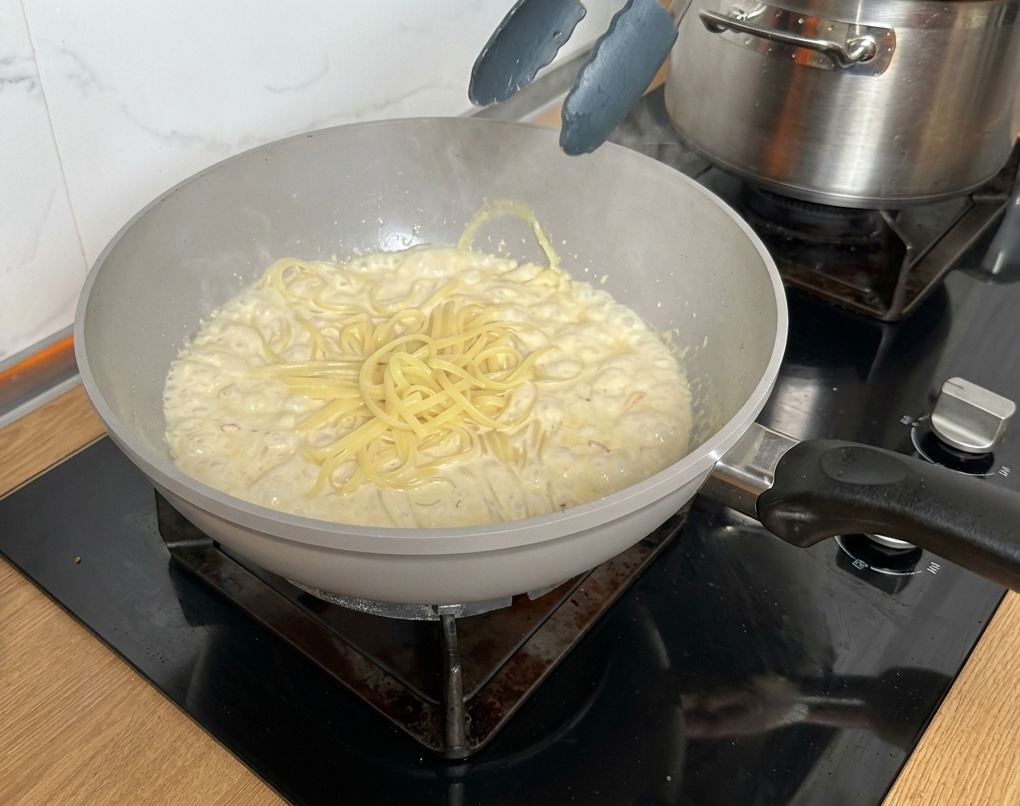 Pinzas metálicas pasando los linguine cocidos directamente a la sartén con la salsa cremosa.
