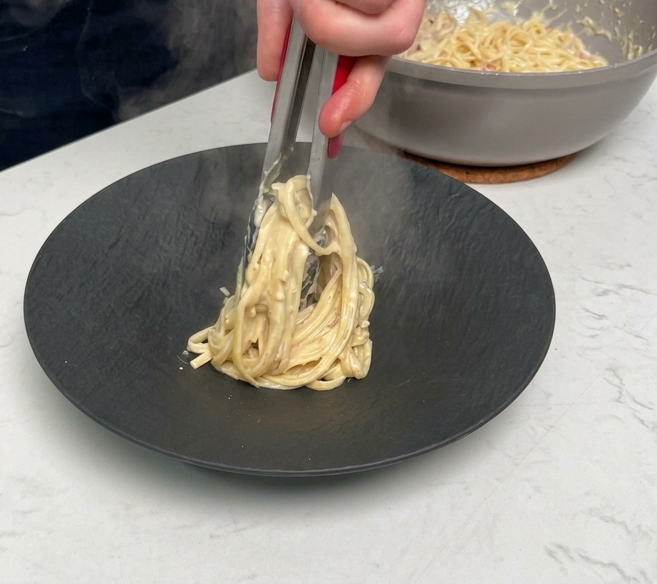 Usando pinzas metálicas para enrollar y servir una ración de pasta cremosa en un plato negro mate.