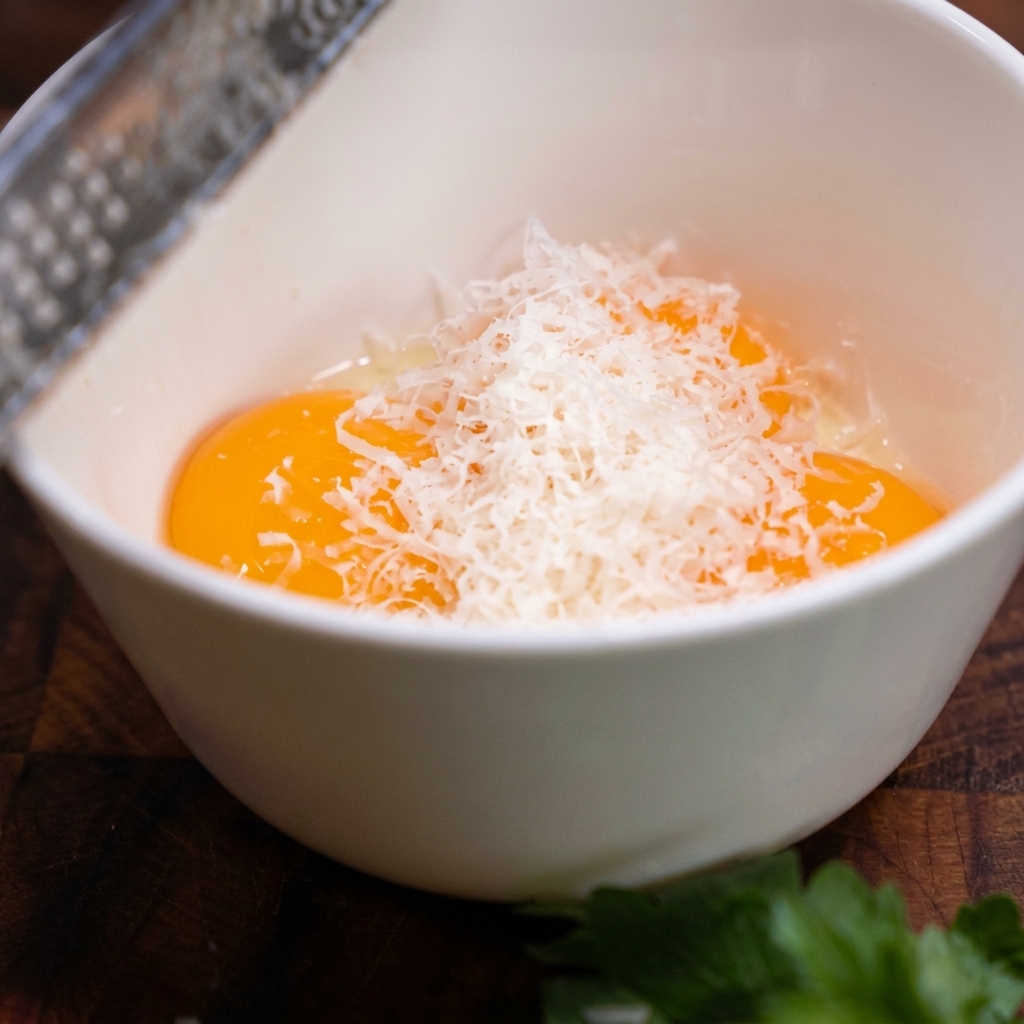 Un rallador de metal rallando finas virutas blancas de queso parmesano en un tazón que contiene tres yemas de huevo crudas.
