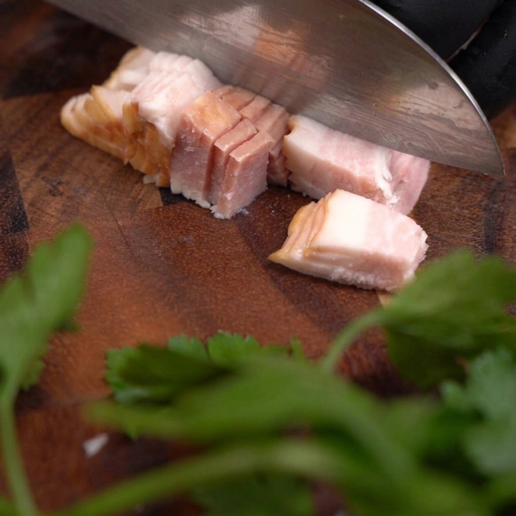 Un cuchillo afilado cortando una tira de tocino crudo para crear cubos pequeños y uniformes sobre una tabla de madera oscura.