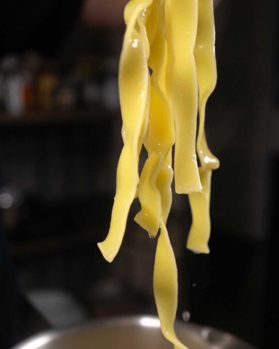 Hilos largos y gruesos de pasta tagliatelle amarilla cocida colgando de unas pinzas, goteando agua.