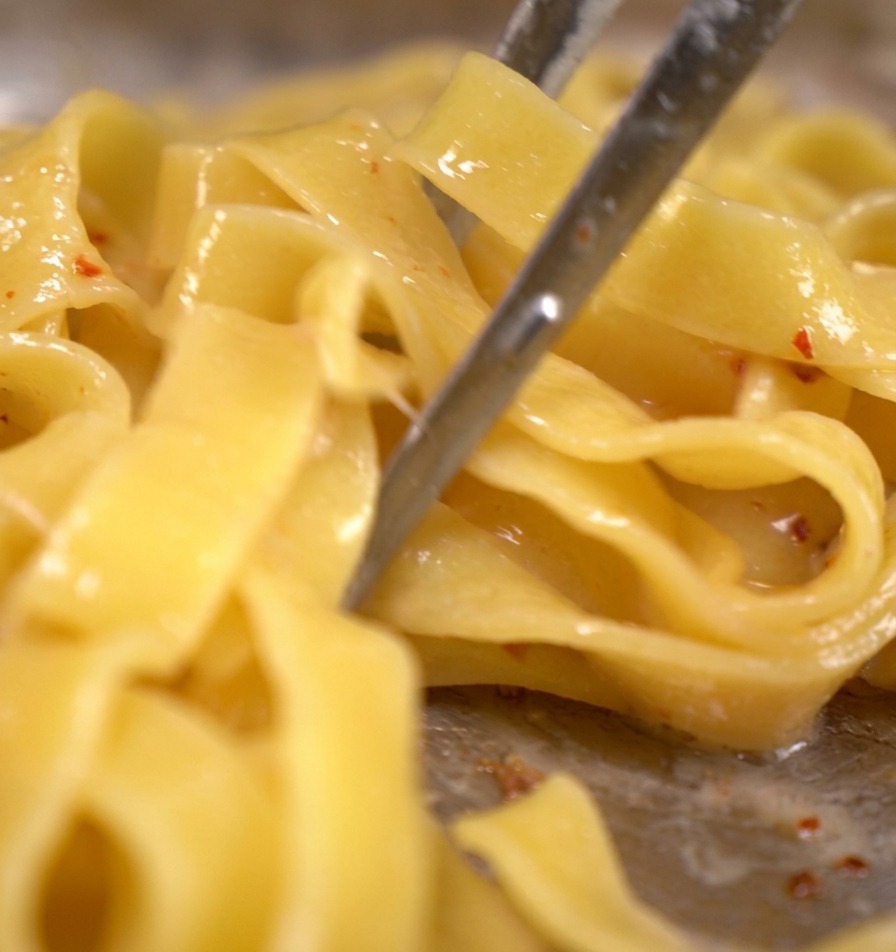 Pasta tagliatelle siendo removida con pinzas de metal en una sartén con tocino picado y agua de cocción de la pasta hirviendo a fuego lento.