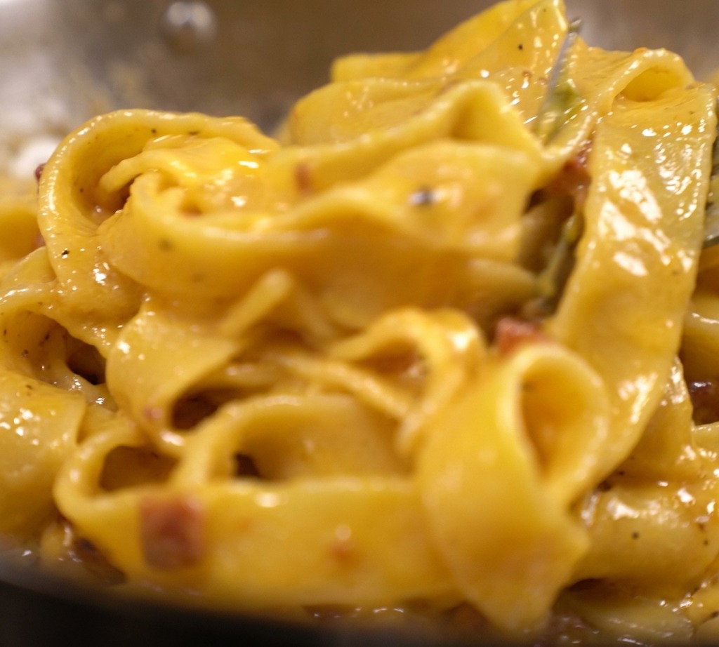 Pasta tagliatelle cubierta con una espesa y vibrante salsa amarilla y cremosa de huevo y queso con trozos visibles de tocino en una sartén.