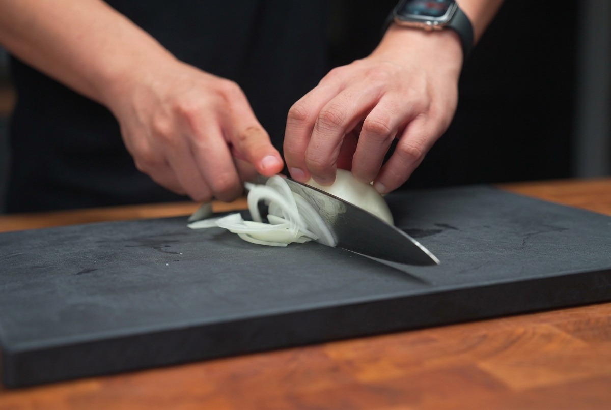 Manos sosteniendo un cuchillo de chef, cortando una cebolla blanca en tiras finas sobre una tabla oscura.