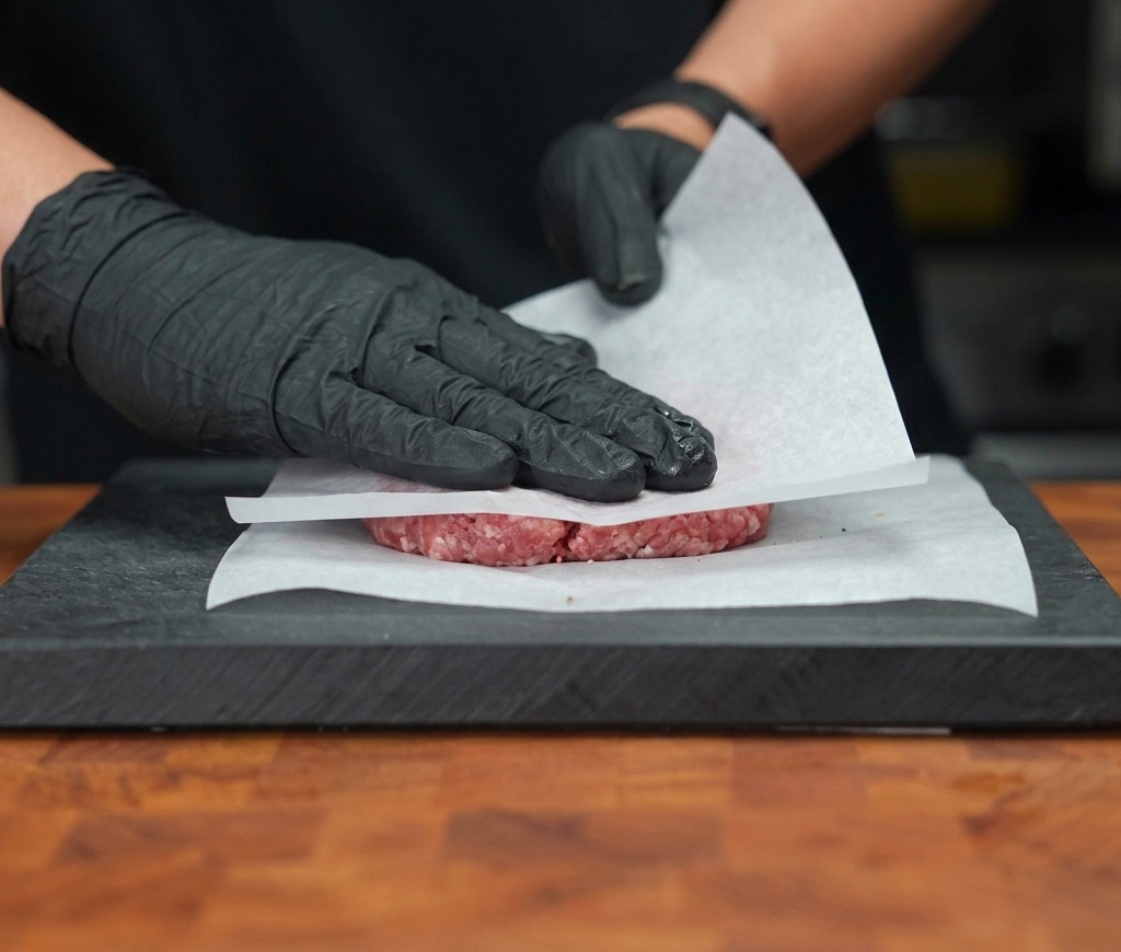 Manos con guantes presionando firmemente un medallón de carne cruda entre dos hojas de papel pergamino.