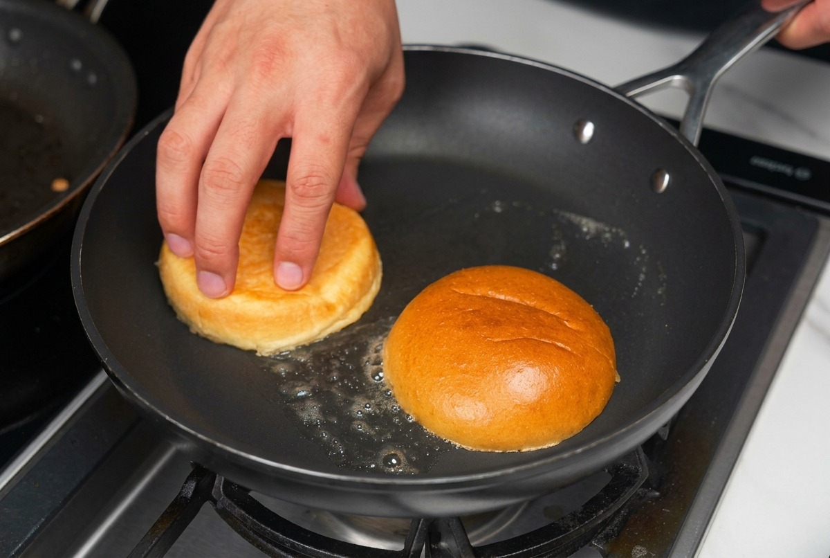 Mitades de pan de hamburguesa tostándose boca abajo en una sartén con mantequilla derretida burbujeante.