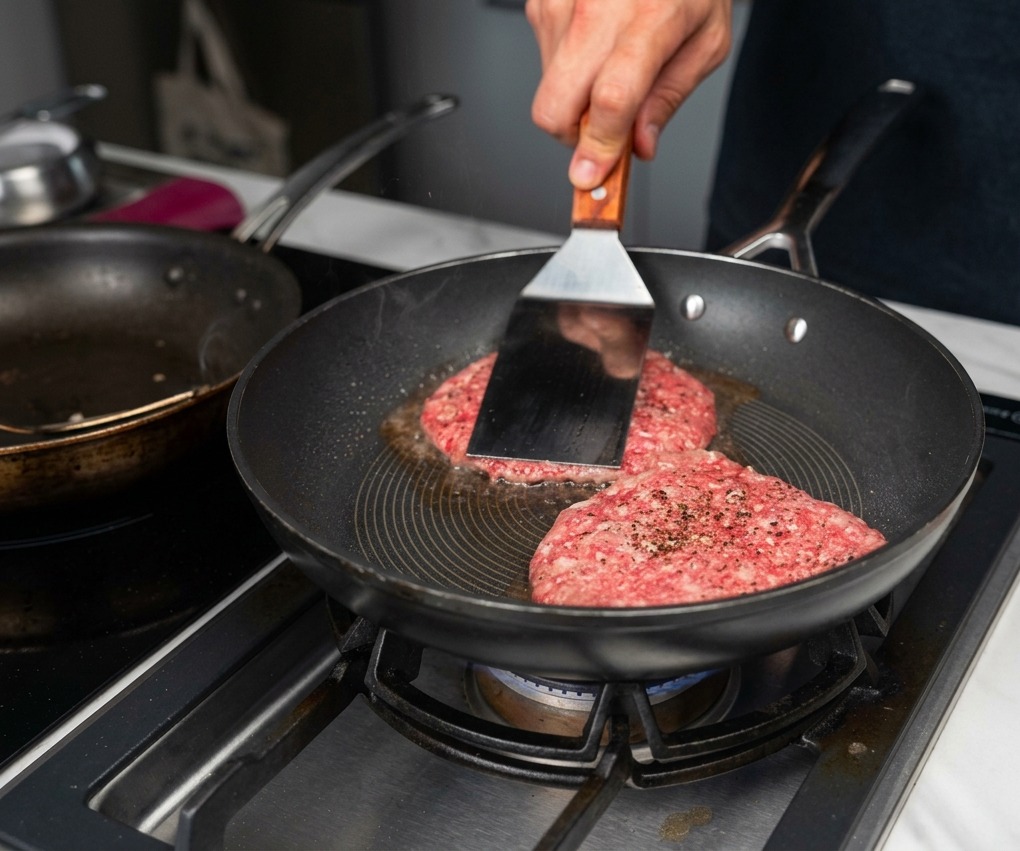 Dos medallones de carne en una sartén caliente mientras una espátula de metal presiona uno de ellos contra la superficie.