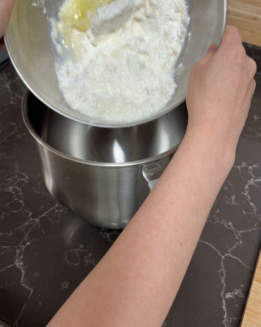 Manos vertiendo una mezcla grumosa de harina e ingredientes húmedos en el tazón vacío de una batidora de pie.