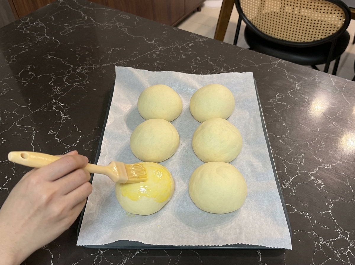 Una mano usando un pincel de repostería para cubrir la parte superior de un pan fermentado con huevo batido brillante.
