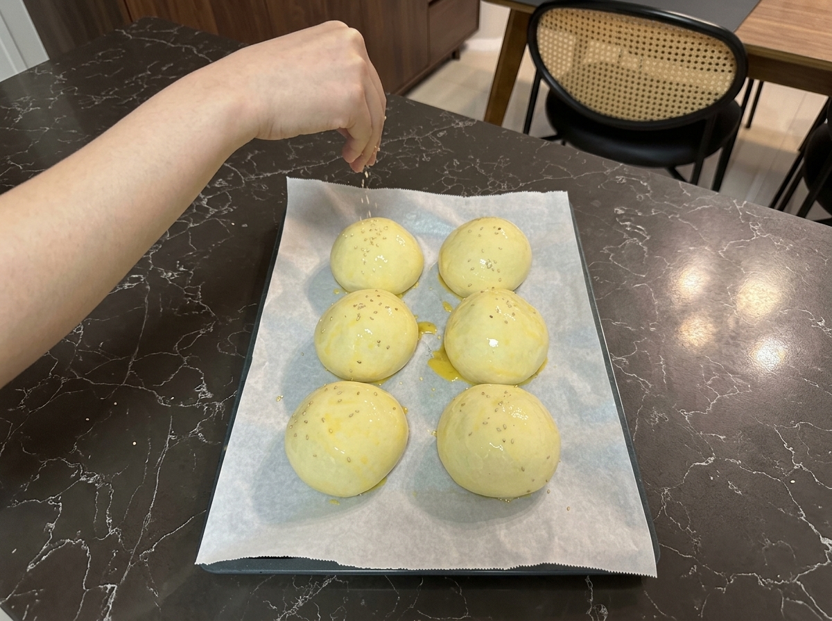 Una mano espolvoreando pequeñas semillas de sésamo blanco sobre las bolas de masa pintadas con huevo en la bandeja.