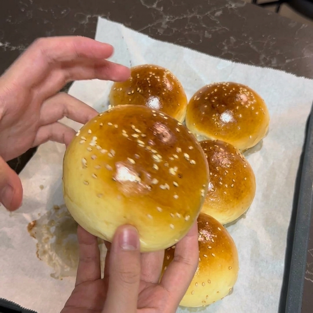 Las manos de una persona sosteniendo delicadamente un pan de hamburguesa recién horneado, dorado y espolvoreado con sésamo.
