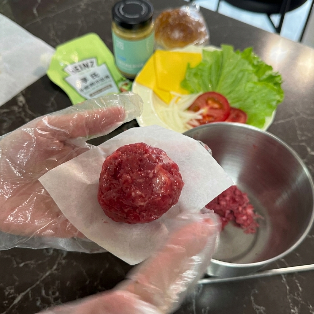 Una mano enguantada sosteniendo una bola redonda de carne picada cruda sobre un pequeño cuadrado de papel vegetal blanco.