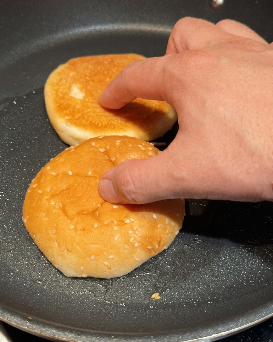 Una mano presionando ligeramente las mitades de un pan de hamburguesa sobre una sartén negra caliente.