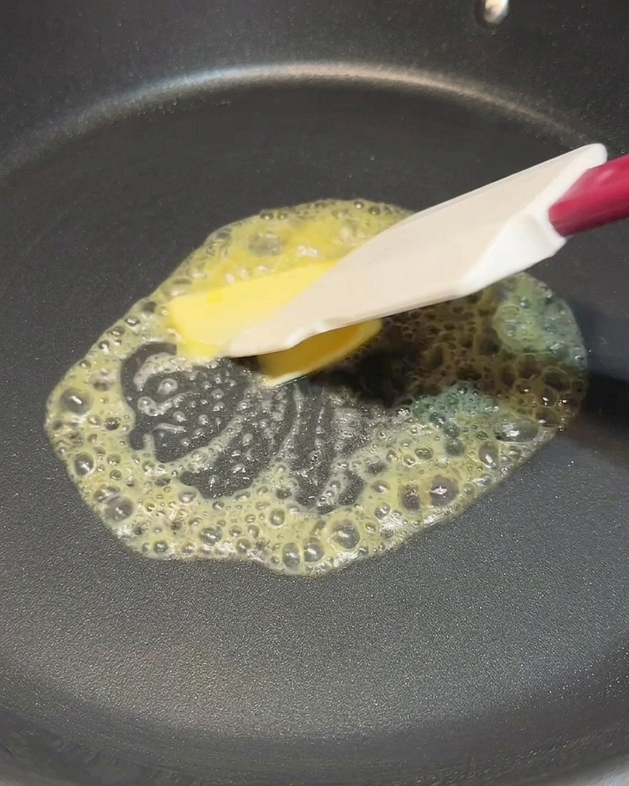 Una espátula blanca esparciendo un cuadro de mantequilla que se derrite en una sartén negra.