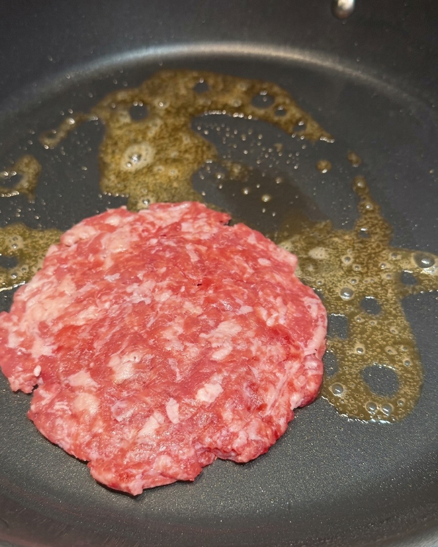 Una hamburguesa de carne cruda y marmoleada cocinándose en mantequilla derretida dentro de una sartén.
