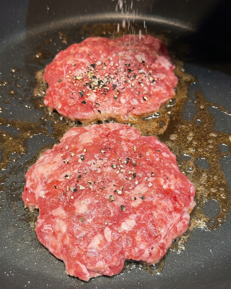 Espolvoreando sal gruesa y pimienta negra sobre dos carnes de hamburguesa en la sartén.
