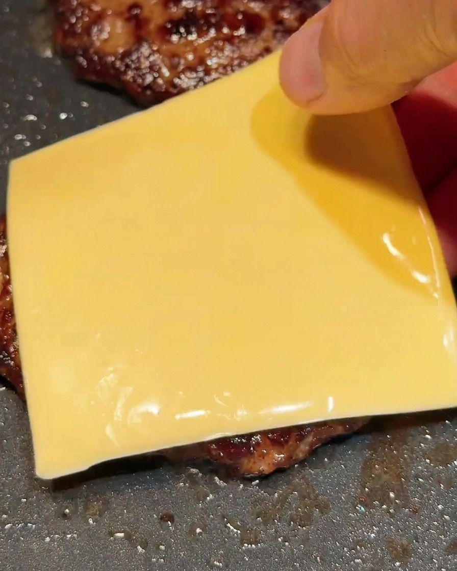 Una mano colocando una rebanada cuadrada de queso amarillo sobre una carne de hamburguesa caliente.