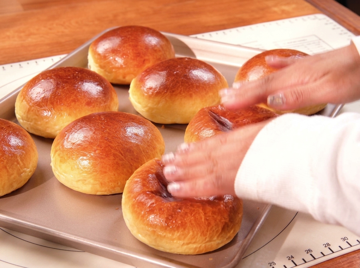 Una mano tocando ligeramente la parte superior de unos panes brioche dorados y brillantes en una bandeja de horno.