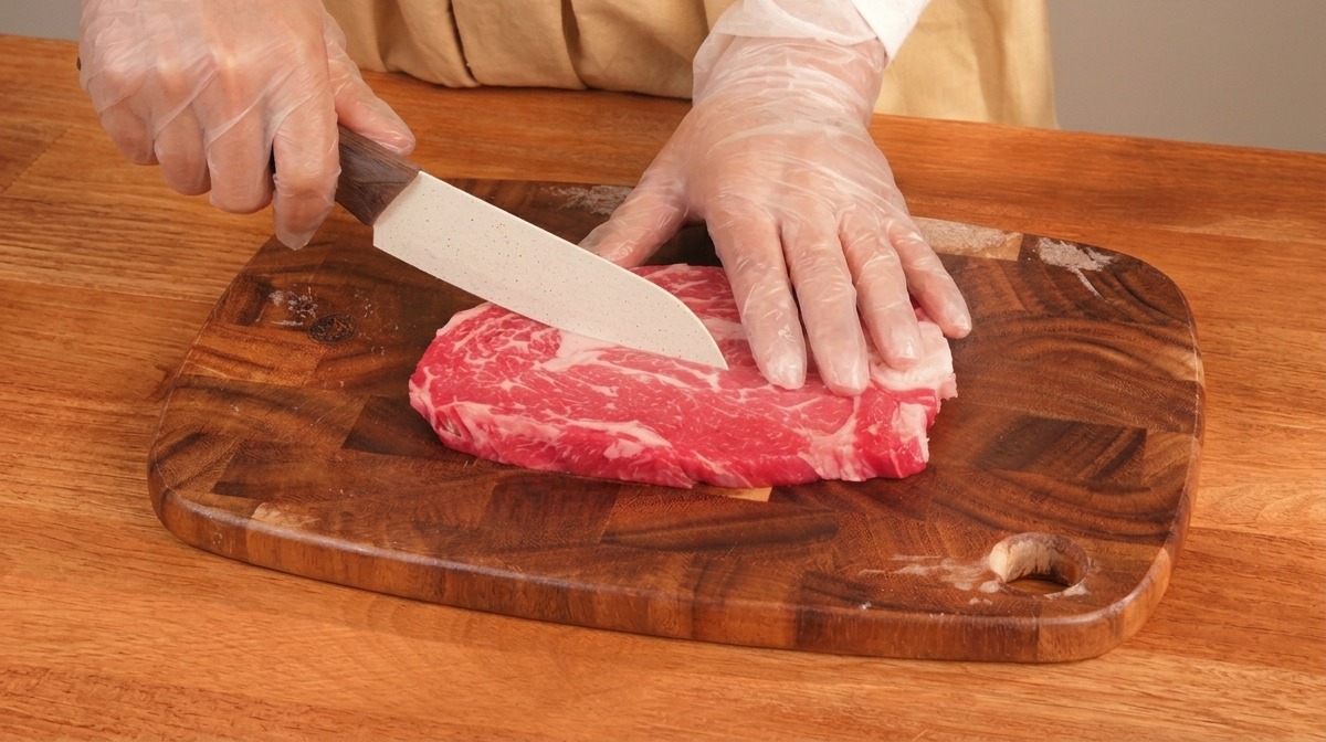 Manos con guantes usando un cuchillo para cortar un trozo grueso de ternera veteada sobre una tabla de madera.