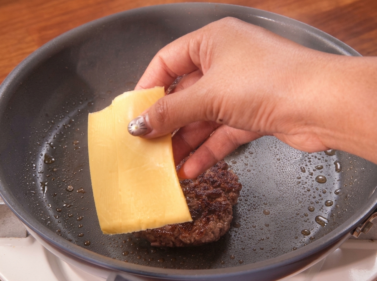 Una mano colocando una loncha de queso amarillo sobre un medallón de carne recién volteado en la sartén.