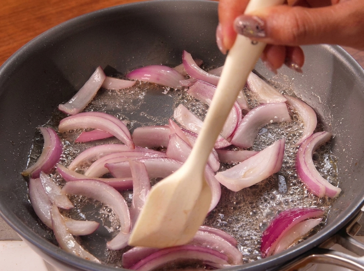 Una espátula removiendo rodajas de cebolla morada mientras se sofríen en una sartén.