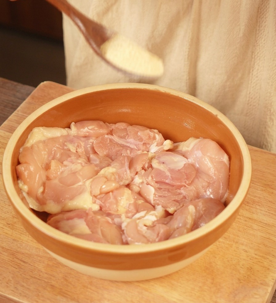 Una cuchara de madera espolvoreando ajo en polvo sobre contramuslos de pollo crudos en un bol de cerámica.
