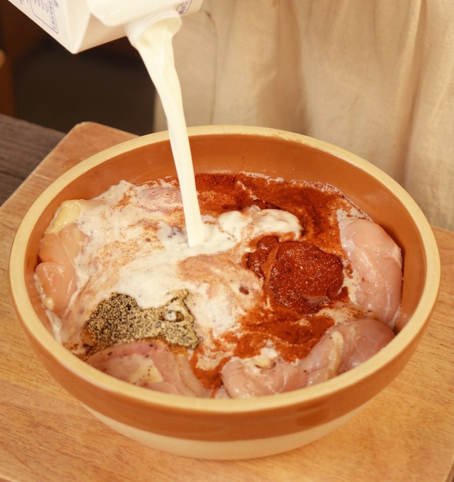 Leche fresca siendo vertida desde un cartón a un bol con pollo crudo sazonado.