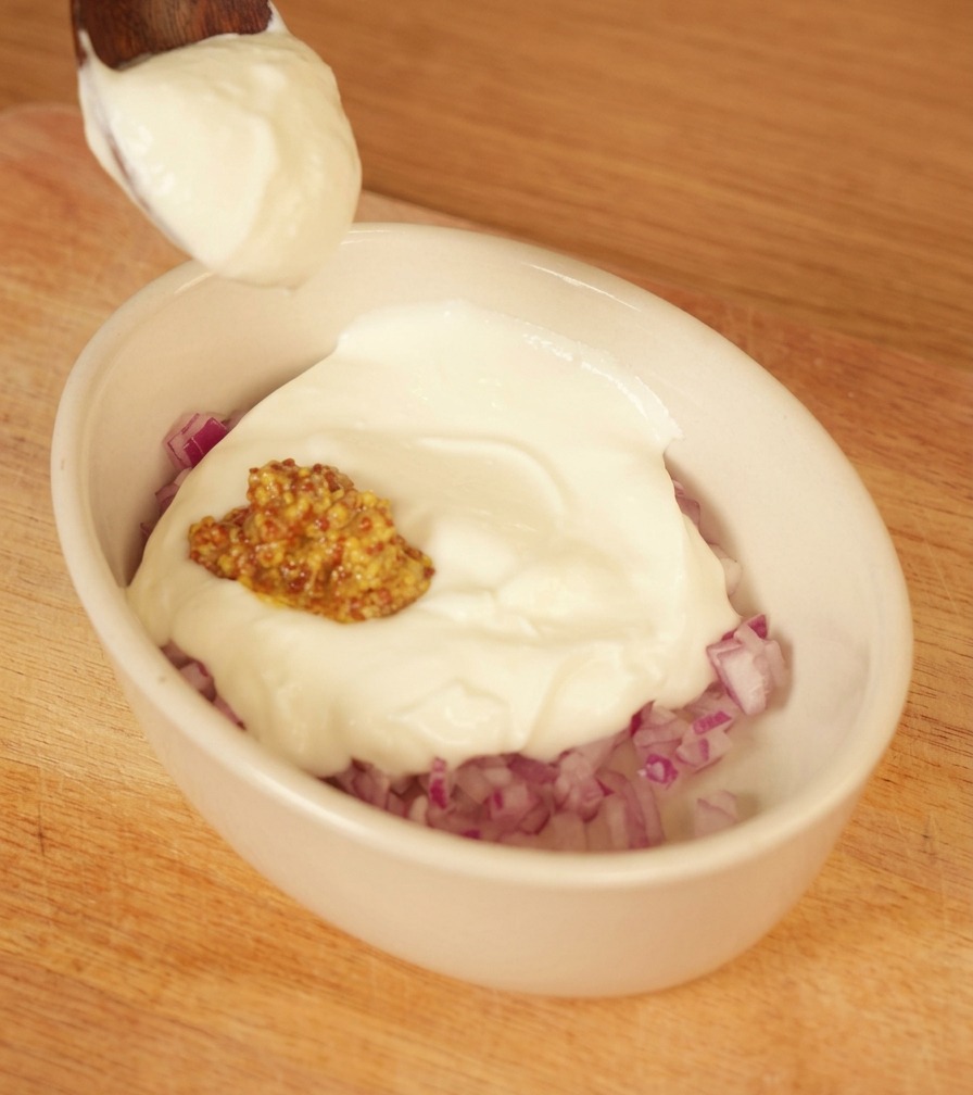 Una cucharada de yogur blanco añadiéndose a un bol con cebolla roja picada y mostaza.