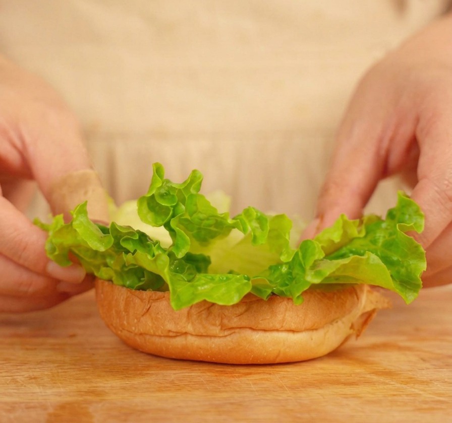 Manos colocando hojas de lechuga sobre la base de un pan de hamburguesa tostado.