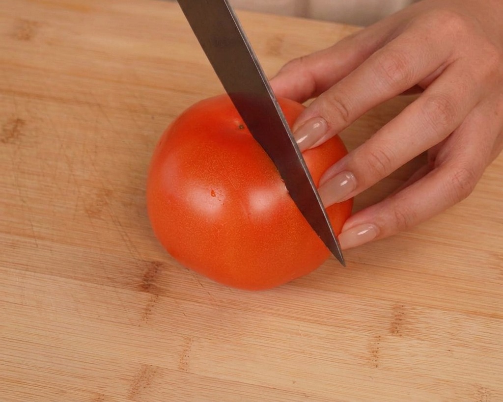 Un cuchillo afilado realizando un corte superficial en forma de cruz sobre un tomate rojo maduro en una tabla de cortar.