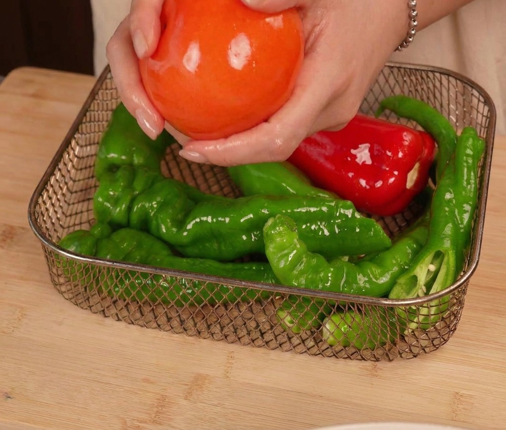 Manos colocando un tomate aceitado y varios chiles verdes y rojos en la cesta de una freidora de aire.