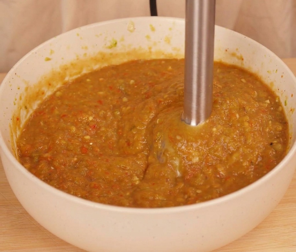 Una batidora de mano procesando chiles y tomate asados hasta obtener una salsa espesa de color naranja amarronado.