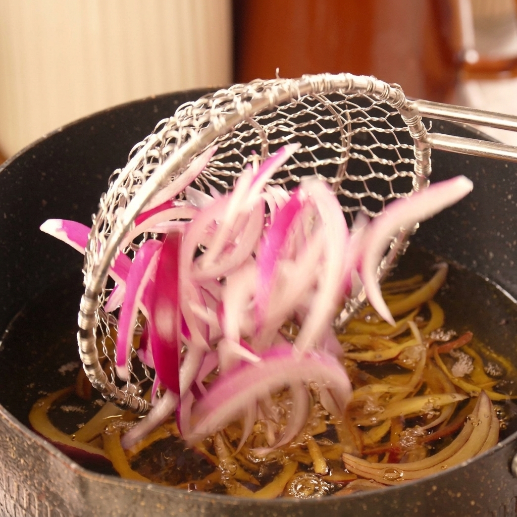 Láminas de cebolla roja introduciéndose en una olla con aceite caliente burbujeante.