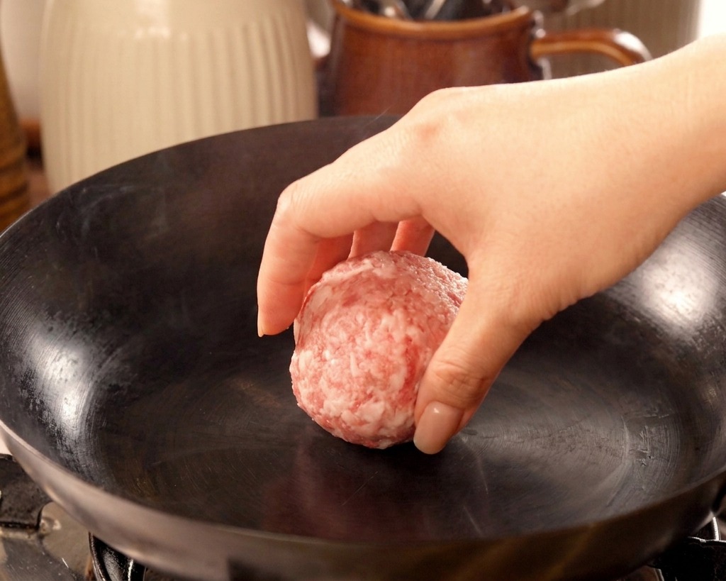 Una mano dejando caer una bola de carne de ternera en un wok de hierro muy caliente sin grasa.