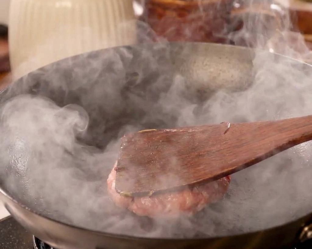 Una espátula de madera presionando fuerte una bola de carne contra la superficie de un wok caliente.