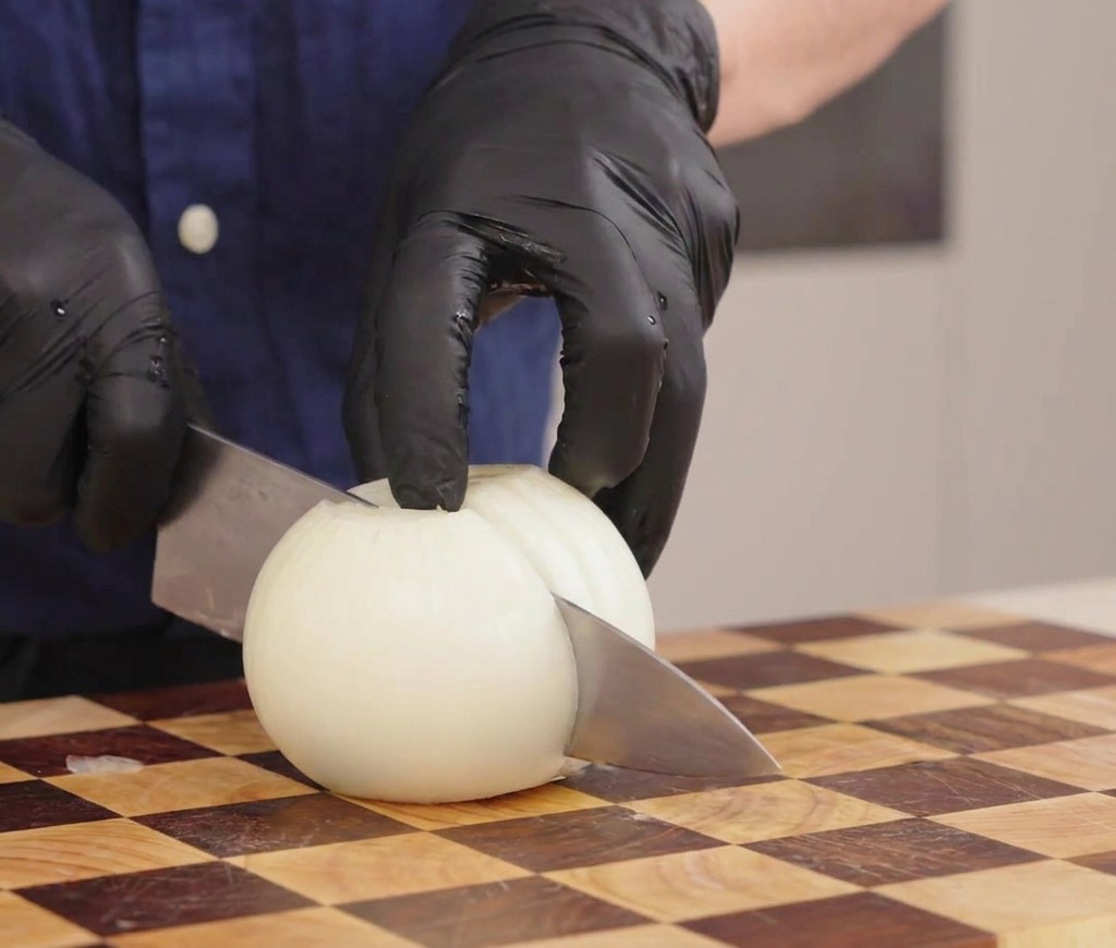 Manos con guantes sujetando un cuchillo para cortar una cebolla blanca en tiras sobre una tabla de madera.