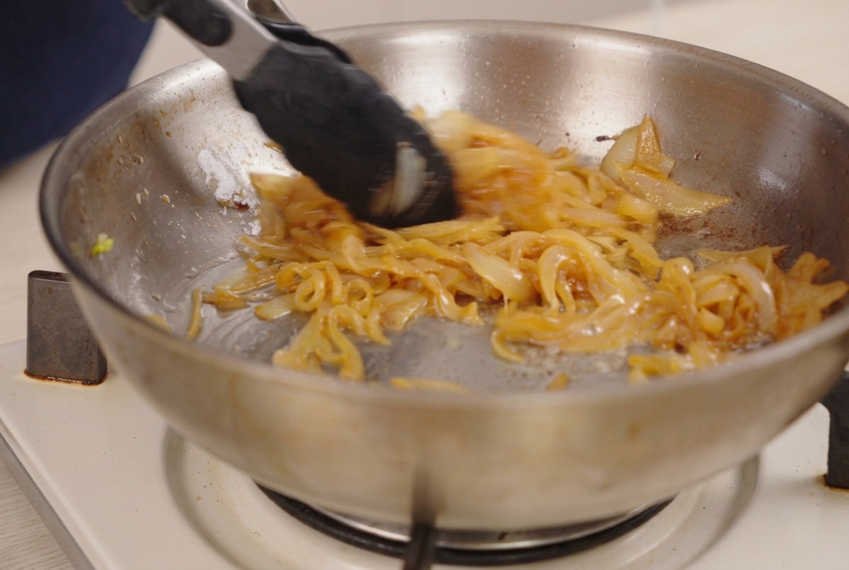 Pinzas removiendo cebollas profundamente doradas y caramelizadas en una sartén.