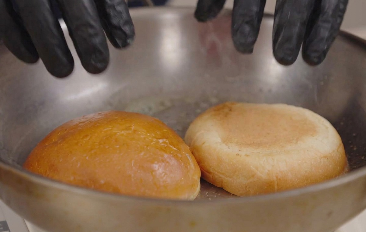Manos con guantes presionando mitades de pan de hamburguesa en una sartén caliente con mantequilla derretida.
