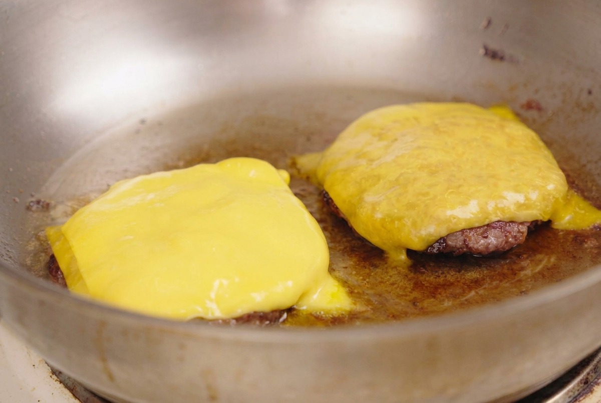 Láminas de queso amarillo derritiéndose sobre dos carnes selladas en una sartén de acero inoxidable.