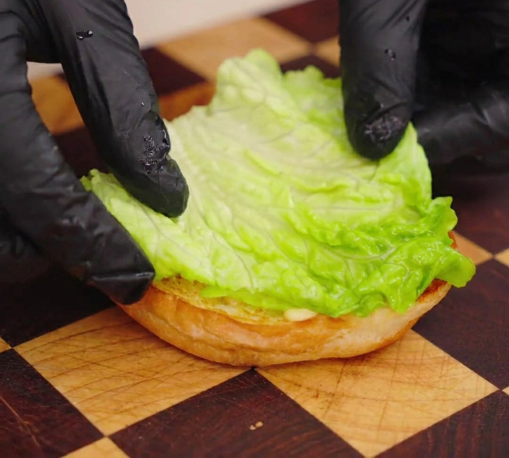 Una persona colocando una hoja de lechuga verde sobre la base tostada de un pan de hamburguesa.