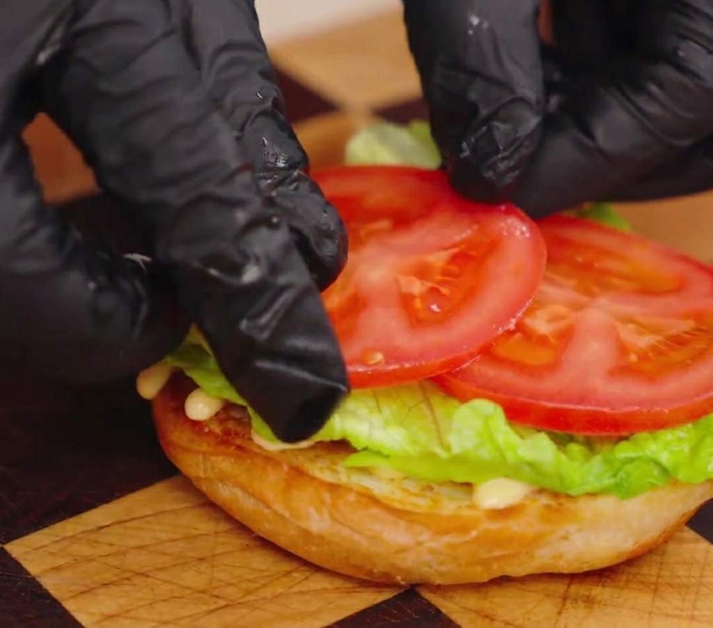 Primer plano de manos acomodando dos rodajas gruesas de tomate rojo sobre la lechuga en el pan.