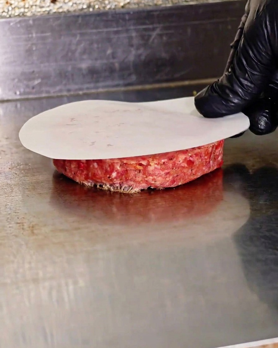 Una mano presionando una hamburguesa cruda con una prensa de metal sobre papel de horno en una plancha caliente.