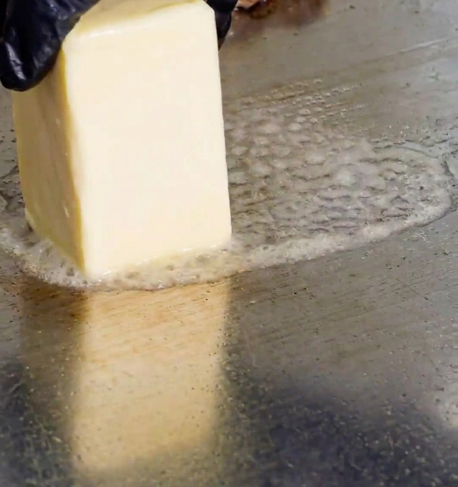 Una mano frotando un bloque de mantequilla sobre la plancha caliente, creando burbujas de grasa derretida.