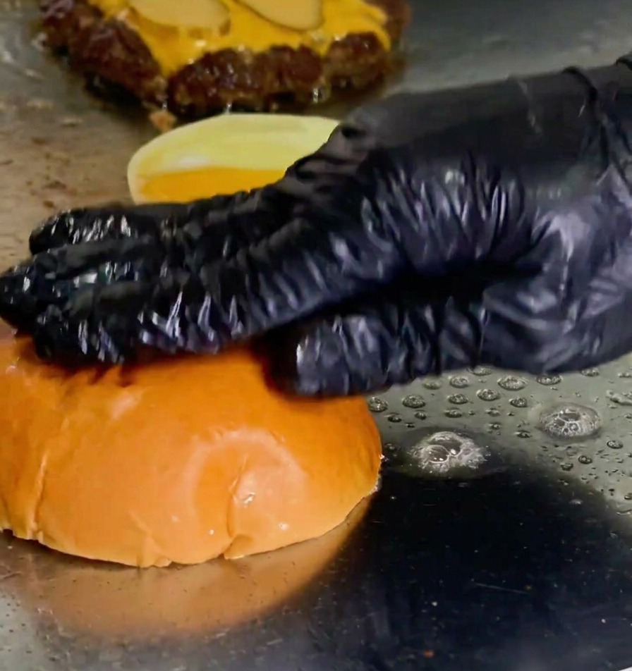 Una mano presionando el pan de hamburguesa sobre la mantequilla derretida en la plancha junto a la carne.