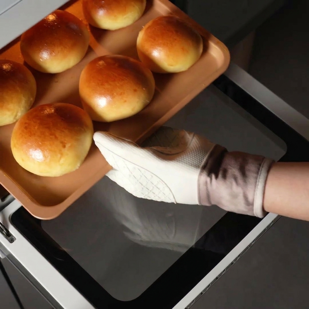 Una mano con guante blanco sacando una bandeja con panecillos de brioche dorados del horno caliente.