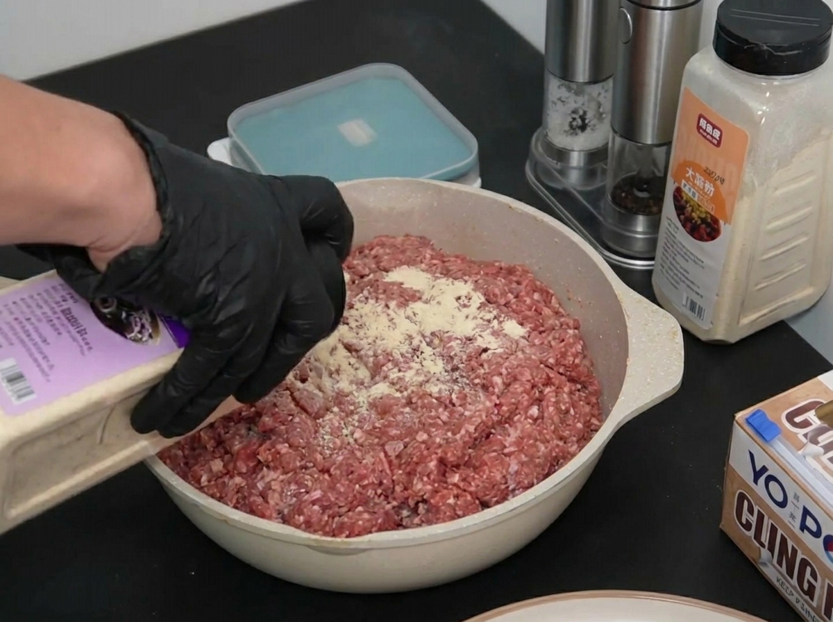 Una mano con guante espolvoreando cebolla en polvo desde un recipiente sobre una bandeja de carne picada cruda.